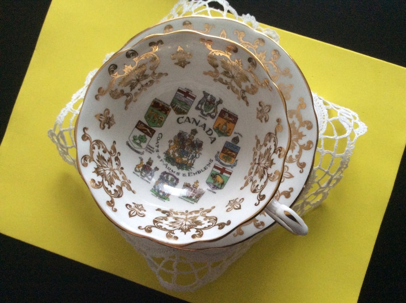 May include: A white teacup and saucer with gold trim and a design of the Canadian Coat of Arms and the provincial coats of arms. The text "CANADA" and "COATS OF ARMS & EMBLEMS" are printed on the cup.