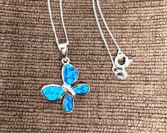 May include: A silver necklace featuring a butterfly pendant with a vibrant blue opal-like inlay. The butterfly design has detailed wings and a delicate silver chain. The clasp is visible at the end of the chain. The necklace is displayed on a textured brown surface.