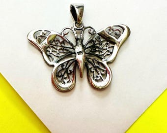 Pode incluir: Pingente de borboleta prateada com intrincados desenhos de asas vazadas. O pingente tem uma argola na parte superior para uma corrente. As asas da borboleta apresentam padrões detalhados, sobre fundo branco e amarelo.