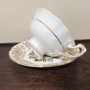 May include: A white teacup and saucer with gold trim. The saucer has a colorful design featuring a coat of arms.