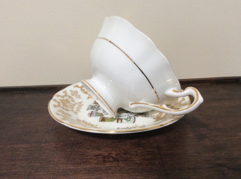 May include: A white teacup and saucer with gold trim. The saucer has a colorful design featuring a coat of arms.
