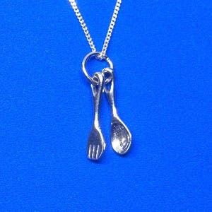 Peut inclure: Un collier argenté avec un petit pendentif en forme de fourchette et de cuillère. Le pendentif est attaché à une chaîne argentée. Le fond est bleu vif.