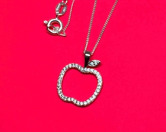 May include: Silver-toned necklace with an apple-shaped pendant. The pendant is outlined with small, clear gemstones. The necklace chain and clasp are also silver. The background is a solid, bright red.