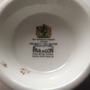 May include: Close-up of the bottom of a white teacup with a gold Paragon logo. The logo reads "By Appointment to Her Majesty the Queen China Potters Paragon Fine Bone China England Reg'd".