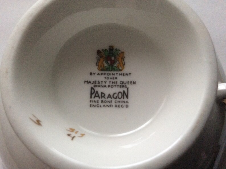 May include: Close-up of the bottom of a white teacup with a gold Paragon logo. The logo reads "By Appointment to Her Majesty the Queen China Potters Paragon Fine Bone China England Reg'd".