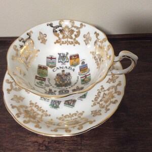 May include: White teacup and saucer with gold trim and a design of the coats of arms of the Canadian provinces. The text "CANADA" is printed in the center of the saucer.