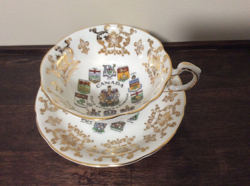 May include: White teacup and saucer with gold trim and a design of the coats of arms of the Canadian provinces. The text "CANADA" is printed in the center of the saucer.