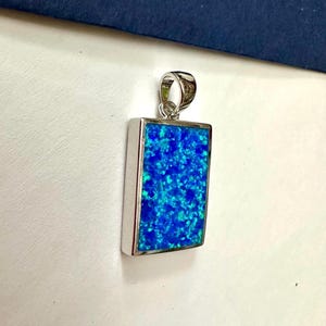 May include: A rectangular pendant with a vibrant blue opal centre set in a silver-coloured frame. The opal has a speckled appearance with shades of blue and turquoise. The pendant has a silver loop for a chain.