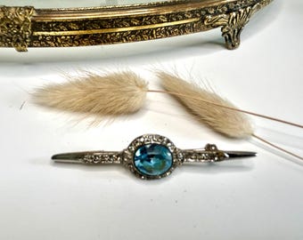 May include: A silver-toned bar brooch featuring a central oval-shaped blue gemstone surrounded by a halo of small clear crystals. The elongated metal pin has a pointed design on both ends and includes a secure clasp mechanism on the reverse side.