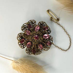 Peut inclure: Broche florale vintage ornée de pierres roses, sertie dans un métal doré filigrané. La broche est dotée d'une fermeture à épingle de sûreté avec une chaîne. Un accessoire décoratif pour vêtements ou accessoires.