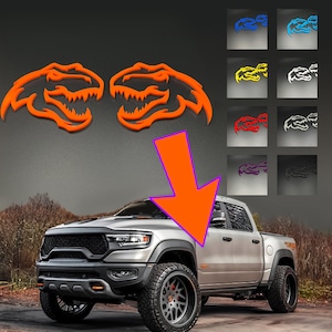 Könnte beinhalten: Ein Paar orangefarbene T-Rex-Kopf-Embleme über einem silbernen Pickup-Truck. Ein großer orangefarbener Pfeil zeigt von den Emblemen auf den vorderen Kotflügel des Trucks. Ein Raster mit acht kleineren Emblem-Varianten in Blau, Gelb, Rot, Lila, Weiß und Schwarz ist abgebildet.