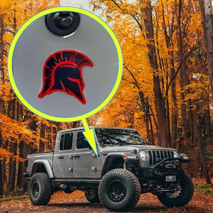 Peut inclure: Un pick-up argenté arborant un emblème de casque spartiate noir et rouge sur le panneau latéral. L'emblème est mis en évidence dans un encart circulaire, montrant un design de casque de guerrier stylisé avec une crête et des lignes angulaires.