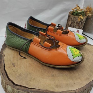 K&ouml;nnte beinhalten: Ein Paar orangefarbene und gr&uuml;ne Lederschuhe mit einem dekorativen Stichdesign an der Spitze. Die Schuhe haben Schn&uuml;rsenkel und ein braunes Lederfutter.