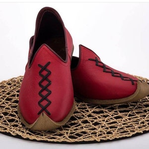 May include: A pair of red leather slippers with black stitching. The slippers have a pointed toe and a flat sole. They are sitting on a woven mat.