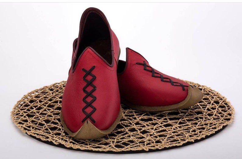 May include: A pair of red leather slippers with black stitching. The slippers have a pointed toe and a flat sole. They are sitting on a woven mat.