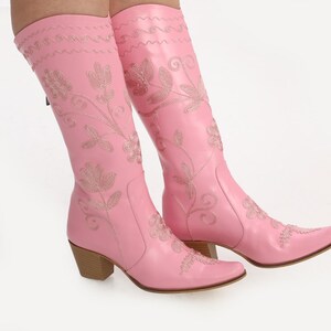 May include: A pair of pink cowboy boots with floral embroidery. The boots have a pointed toe and a block heel.