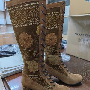 May include: Brown suede knee-high boots with gold embroidery and lace-up fronts. The boots have a block heel and a rounded toe.