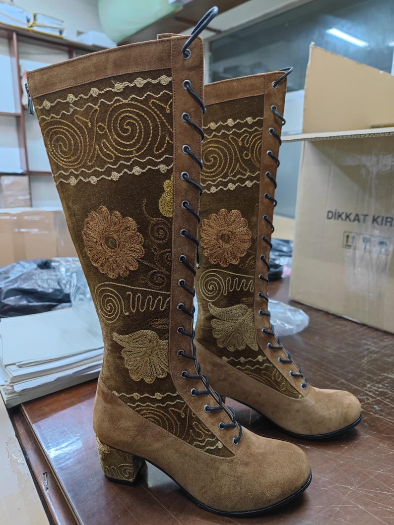 May include: Brown suede knee-high boots with gold embroidery and lace-up fronts. The boots have a block heel and a rounded toe.