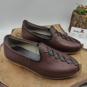 May include: A pair of brown leather slip-on shoes with black stitching and a criss-cross pattern on the front. The shoes have a rounded toe and a flat sole.