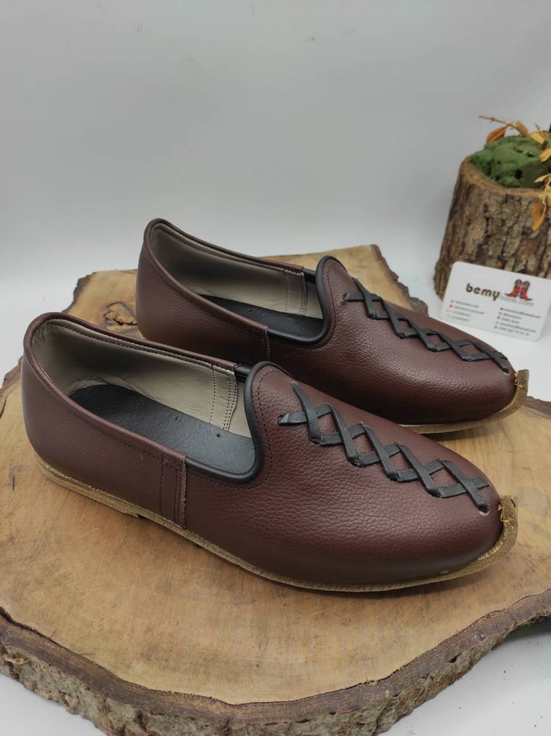 May include: A pair of brown leather slip-on shoes with black stitching and a criss-cross pattern on the front. The shoes have a rounded toe and a flat sole.