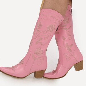 May include: Pink leather cowboy boots with floral embroidery. The boots have a pointed toe and a block heel.