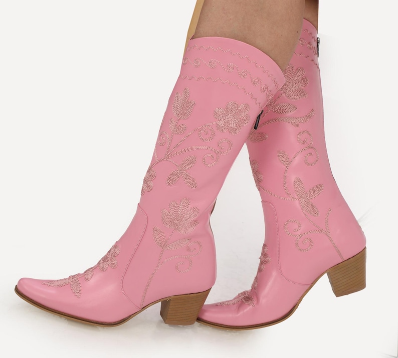 May include: Pink leather cowboy boots with floral embroidery. The boots have a pointed toe and a block heel.