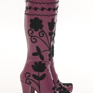 Puede incluir: Un par de botas moradas hasta la rodilla con bordados florales negros y l&iacute;neas onduladas decorativas. Las botas tienen un tac&oacute;n de bloque, punta redondeada y un cierre de cremallera vertical en la parte posterior de la ca&ntilde;a.