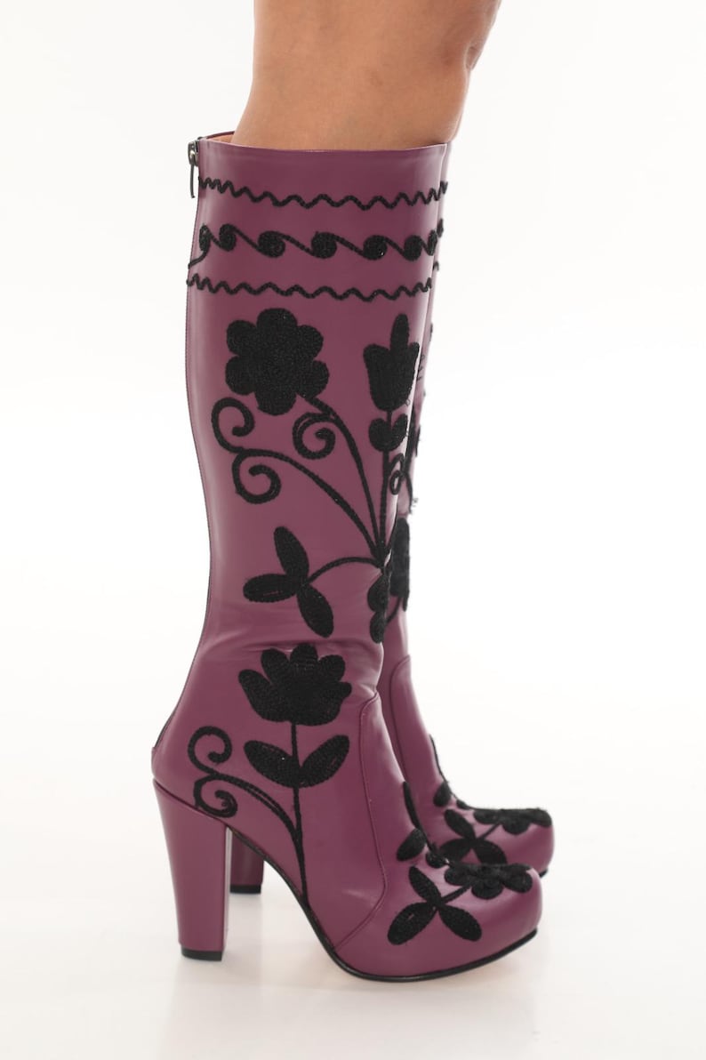 Puede incluir: Un par de botas moradas hasta la rodilla con bordados florales negros y l&iacute;neas onduladas decorativas. Las botas tienen un tac&oacute;n de bloque, punta redondeada y un cierre de cremallera vertical en la parte posterior de la ca&ntilde;a.