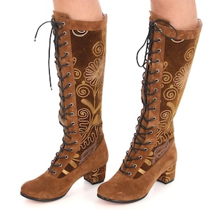 May include: Brown suede knee-high boots with a block heel and gold embroidery. The boots have a lace-up front and a pointed toe.
