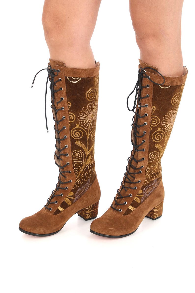 May include: Brown suede knee-high boots with a block heel and gold embroidery. The boots have a lace-up front and a pointed toe.