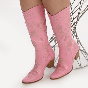 May include: A pair of pink cowboy boots with floral embroidery. The boots have a pointed toe and a block heel.