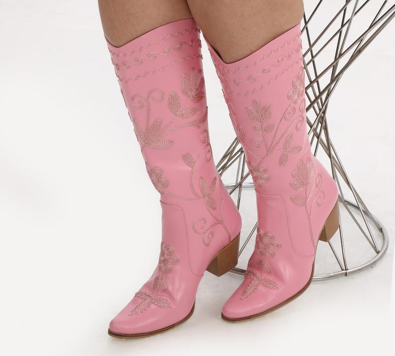 May include: A pair of pink cowboy boots with floral embroidery. The boots have a pointed toe and a block heel.