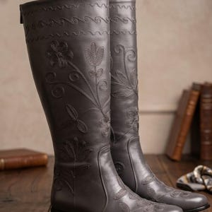 Puede incluir: Botas grises hasta la rodilla con bordados florales. Las botas tienen un patr&oacute;n decorativo cosido en la parte superior y est&aacute;n hechas de un material liso. La palabra "GRAY" est&aacute; impresa en letras negras grandes y en negrita encima de las botas.
