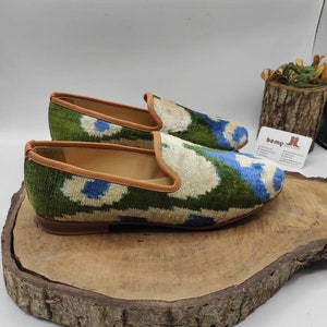 May include: A pair of green, blue, and white patterned velvet loafers with a brown leather trim. The loafers are sitting on a wooden surface.