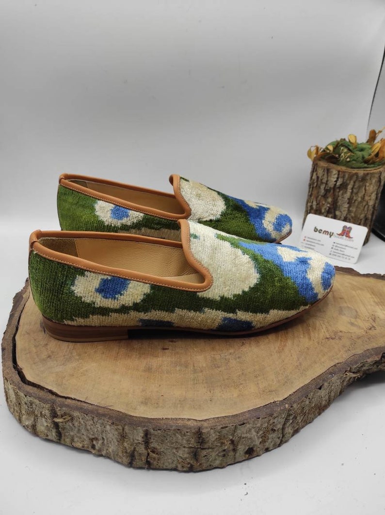 May include: A pair of green, blue, and white patterned velvet loafers with a brown leather trim. The loafers are sitting on a wooden surface.