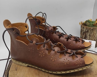 May include: A pair of brown leather sandals with lace-up closures and a floral cut-out design. The sandals are on a wooden surface.