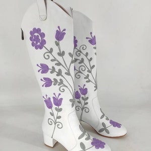 May include: White leather knee-high boots with a floral embroidery design. The flowers are purple and gray.