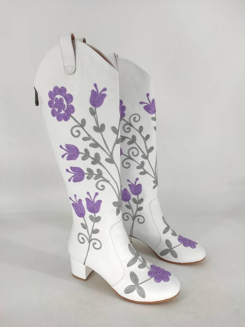 May include: White leather knee-high boots with a floral embroidery design. The flowers are purple and gray.