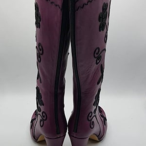 Puede incluir: Un par de botas moradas hasta la rodilla con bordados negros de motivos florales y volutas. Las botas cuentan con una cremallera completa en la parte trasera, un tac&oacute;n de bloque y un forro interior claro visible en la abertura superior.