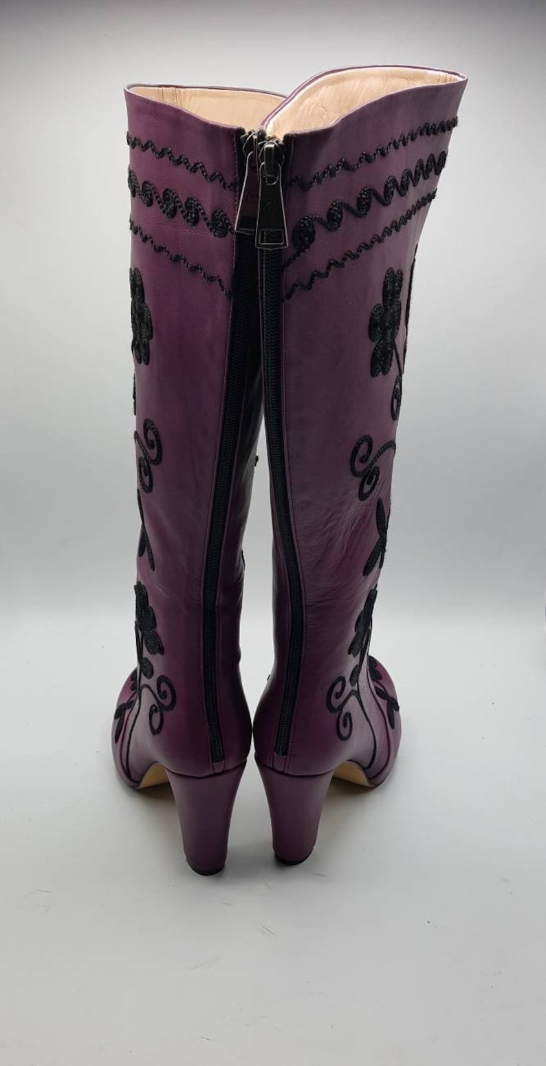 Puede incluir: Un par de botas moradas hasta la rodilla con bordados negros de motivos florales y volutas. Las botas cuentan con una cremallera completa en la parte trasera, un tac&oacute;n de bloque y un forro interior claro visible en la abertura superior.