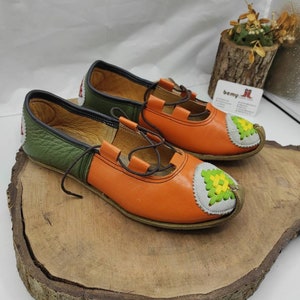 K&ouml;nnte beinhalten: Ein Paar orangefarbene und gr&uuml;ne Lederschuhe mit einem dekorativen Stichdesign an der Spitze. Die Schuhe haben einen Schn&uuml;rverschluss und eine braune Ledersohle.