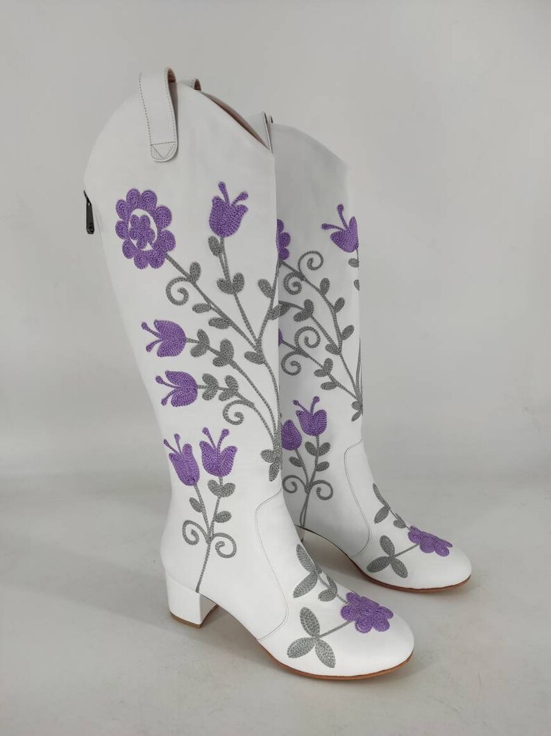 May include: White leather knee-high boots with a block heel and purple floral embroidery.
