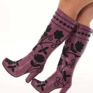 Puede incluir: Botas altas de color morado con bordados florales negros y un patr&oacute;n de l&iacute;neas onduladas en la parte superior. Estas botas cuentan con un tac&oacute;n de bloque, punta redondeada y cierre de cremallera lateral, con costuras negras sobre la base morada.