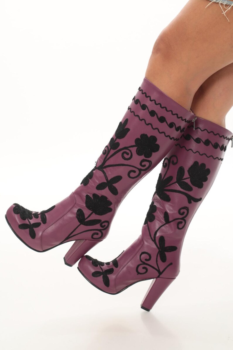 Puede incluir: Botas altas de color morado con bordados florales negros y un patr&oacute;n de l&iacute;neas onduladas en la parte superior. Estas botas cuentan con un tac&oacute;n de bloque, punta redondeada y cierre de cremallera lateral, con costuras negras sobre la base morada.