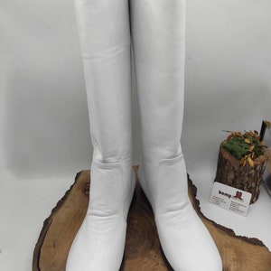 Puede incluir: Un par de botas blancas de cuero de ca&ntilde;a alta con punta redonda y tac&oacute;n bajo. Las botas se muestran sobre una superficie de madera.