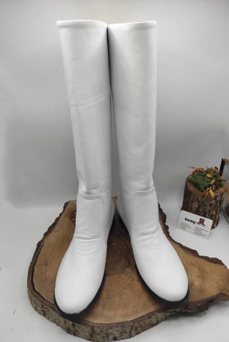 Puede incluir: Un par de botas blancas de cuero de ca&ntilde;a alta con punta redonda y tac&oacute;n bajo. Las botas se muestran sobre una superficie de madera.