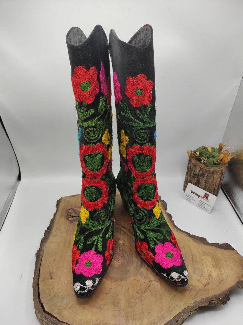 May include: A pair of black knee-high boots with colorful floral embroidery. The boots have a pointed toe and a low heel.
