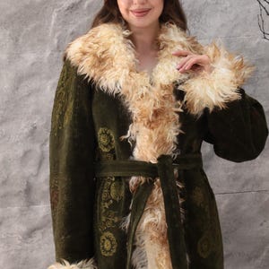Puede incluir: Abrigo largo verde oliva con cuello y puños de piel sintética. El abrigo presenta un cinturón a juego y patrones florales dorados. La piel sintética es de color marrón claro, creando un contraste con la tela verde oscuro.