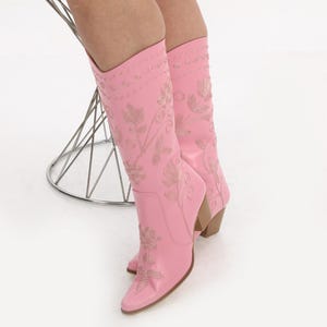 May include: A pair of pink cowboy boots with floral embroidery. The boots have a pointed toe and a high heel.