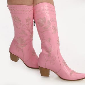 May include: Pink leather cowboy boots with floral embroidery. The boots have a pointed toe and a block heel.